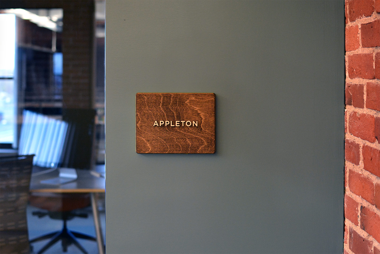 Architectural room identifying sign for a room named Appleton, made out of stained wood, installed on slate colored drywall with exposed brick and glass walls showing at the edges.